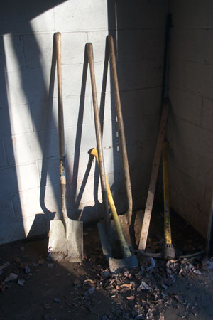 Three shovels, an axe, and a pick-axe, lined up against a cinder block wall