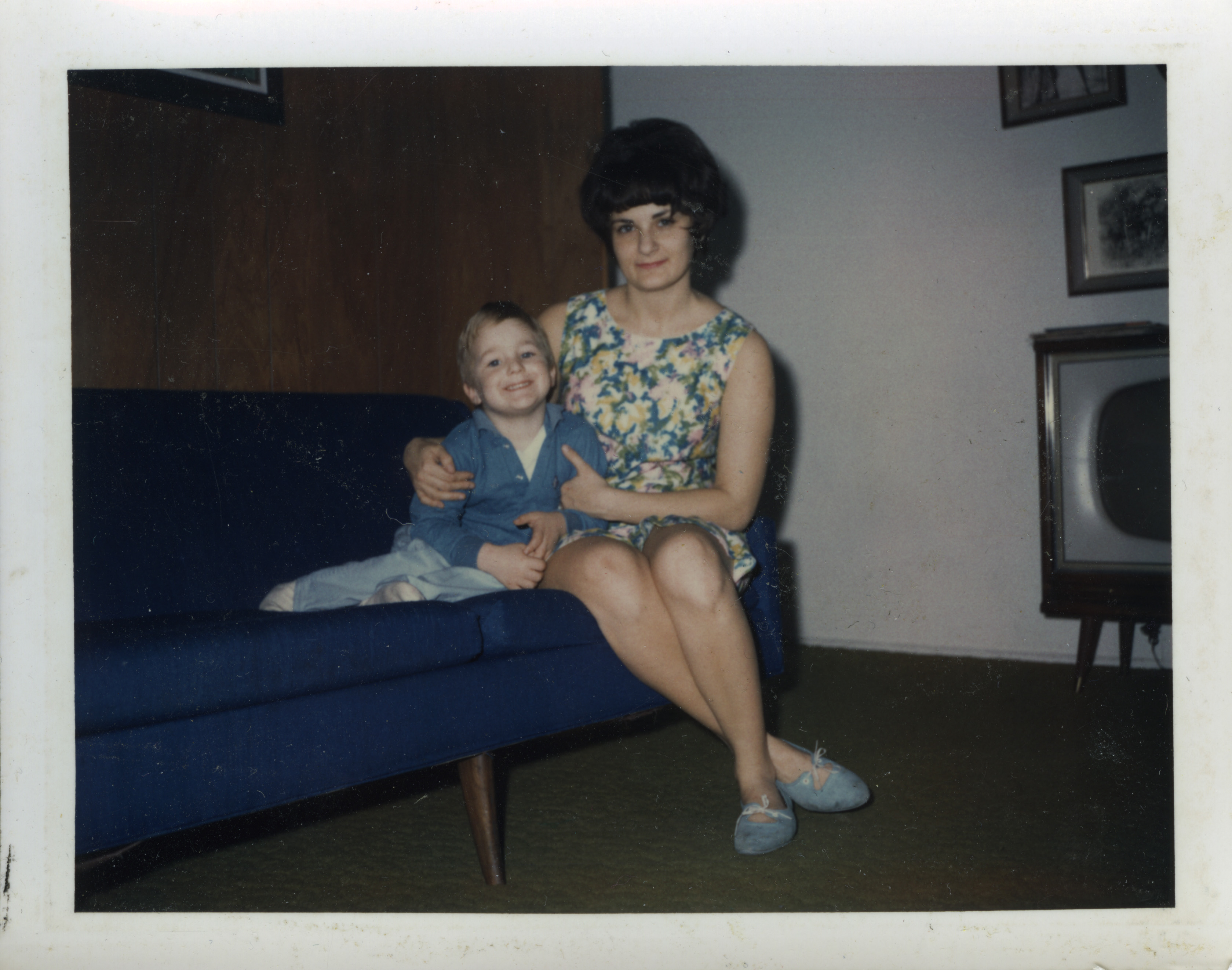 A young boy and an attractive woman in a flowered dress sit on a solid blue couch in front of a paneled wall. The woman has her arms around the boy. The photograph is a color Polaroid shot on peel apart film.